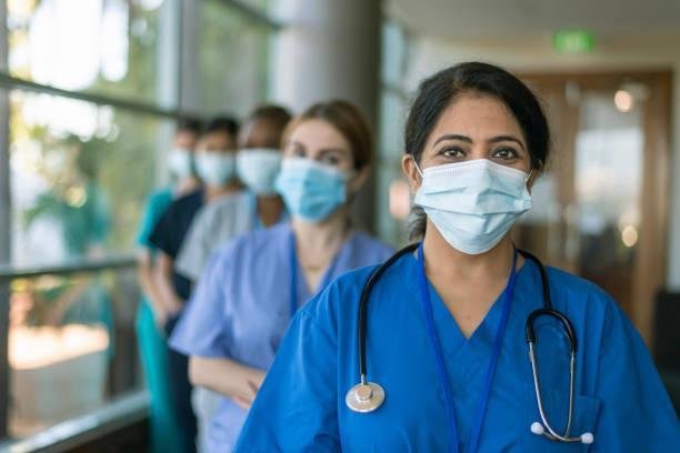 A multi-ethnic team of medical workers wearing scrubs and protective face masks look directly at the camera while posing in a line in a hospital corridor